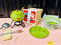 Full view of fondue set components: green fondue pot with white floral decoration, black metal stand, silver fuel burner, four fondue forks in plastic, green divided plate, vintage original box and instruction sheet.