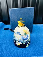 Porcelain honey pot with floral decorations and 3D bee lid seen from front, original box in background.