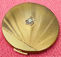Top view of gold-tone powder compact with textured pattern and RCAF emblem on lid, placed on a pink fabric surface.