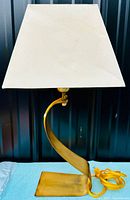 Front view of vintage brass table lamp with curved abstract base and large white trapezoidal fabric lampshade showing dirt and stains.