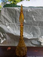 Full frontal view of the tall amber glass genie bottle with a pointed hobnail stopper and dimpled glass body on a wooden surface against a light backdrop outdoors.