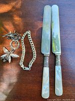 Photo showing two sterling silver cutting knives with mother of pearl handles, a curb link chain bracelet, and three brooch pins on a wooden surface.