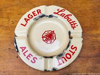 Antique round enameled metal ashtray with Labatt's branding showing multiple chips and worn spots on the enamel surface. Red lettering around edge and red Labatt's logo in center.