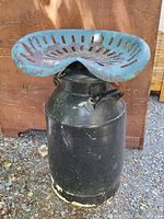Black antique metal milk can with black paint showing wear and chipped paint at the base. Cast iron tractor seat on top painted blue with rust and wear visible.