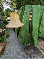 Full view of the Art Deco wood and metal bridge lamp standing outdoors on gravel with a green tarp background. Shows the curved arm and cream fabric shade with decorative trim.