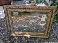 Photo showing close-up view of the rectangular antique mirror with ornate textured gold-tone frame placed outdoors on gravel ground.