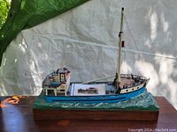 Photo of hand carved fishing trawler model La Levisienne showing the full side profile mounted on a wooden base with ocean-like texture.