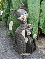 Two wooden folk art cat figurines standing on gravel with green tarps in background. Larger figure is about 40 inches tall with gray and white stripes and blue eyes, smaller figure sitting beside it with matching paint and facial details.