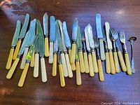 Wide view showing the full assortment laid out on a wooden surface including knives, forks, and a spoon. Handles are mostly cream or yellow celluloid or Bakelite.