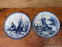 Pair of antique Delft blue and white ceramic wall plates featuring Dutch country scenes with floral borders, placed on wooden surface.