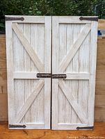 Full front view of the pair of whitewashed wooden wall hanger doors with metal hardware and latch, shown against wooden support.