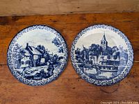 Pair of vintage Delft blue and white wall plates displayed on wood surface showing front face with detailed Dutch scenes and floral borders.