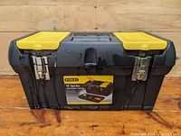 Front view of Stanley plastic toolbox with metal latches and yellow compartments on top, showing label and handle.