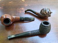 Three vintage tobacco pipes shown on wooden surface: a smooth wood pipe with metal band, a textured rustic pipe, and a carved figure-head pipe.