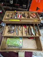 Top view of open beige Plano tackle box showing double fold-out trays filled with assorted fishing lures including metal spoons, plastic and wooden lures with hooks.