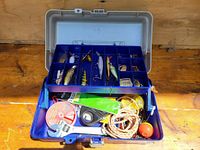 Photo of open fishing tackle box showing upper tray with various vintage fishing lures and lower compartment with rope, fishing line, float, tape, and other accessories.
