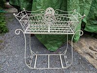 Front view of white vintage metal wire plant stand with ornate floral decoration and scroll legs on gravel surface outdoors