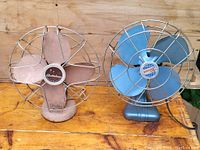 Both vintage metal fans side by side on wooden surface, showing size, color, and protective cages.