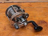 Side view of circular metal fishing reel with thin wire line and black handle knobs on wooden surface.