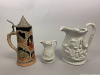 Photo shows a vintage German beer stein with a pewter lid and two Portmeirion Parian Ware white ceramic pitchers with embossed detailed scenes.