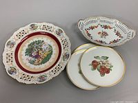 Wide view of all items in the lot showing large pierced plate with classical scene, oval floral bowl, and three smaller fruit motif plates