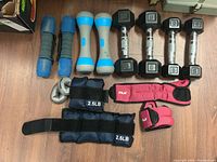 Overview photo showing all items in the lot including dumbbells, wrist supports, and weighted straps laid out on wood floor.