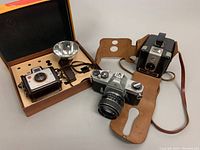 Wide view showing Pentax Spotmatic SPll camera with leather case, Kodak Brownie Holiday Flash Camera with box and attached flash, and Kodak Browne Hawkeye Camera Flash with strap.