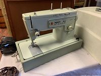 Full front and side view of the pale green Singer Zig Zag sewing machine on a white surface with case partially visible.