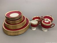 Full set of stacked plates, teacups with saucers, creamer, sugar bowl with lid shown with the same red and gold decorative pattern.