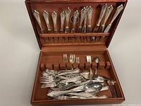 Open wooden chest showing silver plated knives, forks, and spoons arranged inside, handles with ornate classical design.