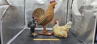 Three rooster figurines displayed together showing relative size and detail, including wood, ceramic, and plaster.