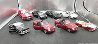 Eight diecast model cars arranged on a reflective surface with a silver backdrop. Various styles and colors including convertibles and coupes. Doors open on some cars showing interiors.