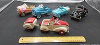 Eight miniature diecast pedal cars arranged on a black surface with a ruler in front for scale. Cars include a white and red convertible with trailer, tan with wooden trailer, blue, turquoise convertible style, and black vintage style pedal cars.