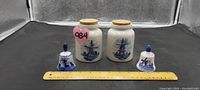Two Delft ceramic jars with cork lids and two small Delft ceramic bells with blue windmill design, placed next to a ruler for size reference.