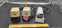 Front view of three electric pencil sharpeners with attached power cords placed behind a wooden ruler for size reference, showing two cream-colored models and one white model.
