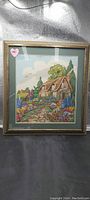 Framed needlepoint picture showing a cottage and colorful garden with flowers, in a wooden frame with green matting.