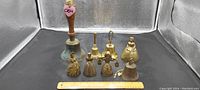 Group of eight brass bells including four Victorian lady bells, one wood handle bell, two dinner bells, and one liberty bell along with a ruler for scale.