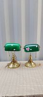 Pair of green glass bankers lamps with polished brass bases and pull chain switches, photographed outdoors on fabric surface.