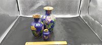 Three blue cloisonné vases displayed together with ruler to indicate size. Vases in 10, 8, and 4 inch approximate heights. Blue background with multicolored floral enamel work.