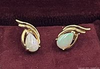 Front view of pear-shaped opal stud earrings in yellow gold swirl setting on burgundy background