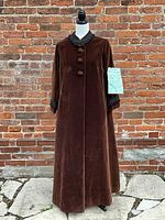 Front view of the full-length brown velvet cape showing silhouette and button closure against brick background.