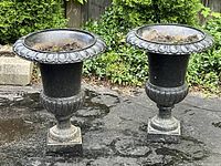 Two black painted cast iron urns side by side on asphalt with garden foliage behind