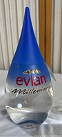 Full view of clear Evian Millennium water bottle with blue conical cap, showing the tapering shape and branding.