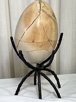Large polished septarian stone egg mounted on a black metal stand with spider-like legs, showing natural beige and tan fracture pattern.