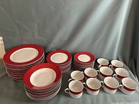 Full display showing stacks of dinner plates with red rims, smaller plates, soup bowls, tea cups and saucers laid out on gray fabric background.