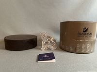 Photo showing the Swarovski crystal grizzly bear figurine next to its original round brown Swarovski Silver Crystal box with foam insert and certificate booklet.