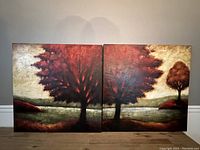 Two square canvas paintings side by side featuring red trees and landscape scene; shot includes full view of art pieces on neutral wall