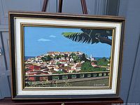 Framed oil on board painting showing full view of a hillside town with red-roofed buildings and greenery under a bright blue sky.