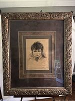 Full view of the framed sketch hung on wall, showing ornate carved wooden frame and large brown double mat surrounding the central drawing of a girl.