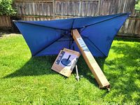 Photo showing the large blue umbrella opened on grass with the boxed umbrella stand and umbrella pole beside it.